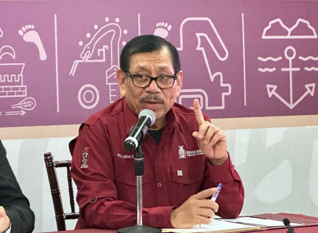 Gerardo Mérida Sánchez con camisa guinda institucional de la Secretaría de Seguridad, gesticulando y apuntando con el dedo durante una conferencia de prensa frente a un fondo morado con glifos.