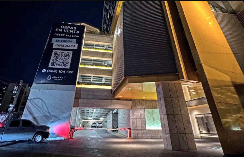 Entrada del estacionamiento del edificio Adamant Grand en Tijuana por la noche, con la pluma de control de acceso baja. Se aprecia un anuncio de venta de departamentos de Adamant Grand.