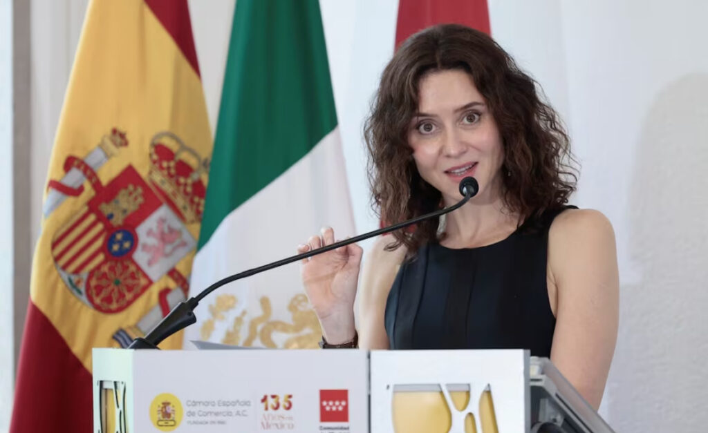 Isabel Díaz Ayuso, presidenta de la Comunidad de Madrid, dando un discurso en un podio con la bandera de España y la bandera de México de fondo.