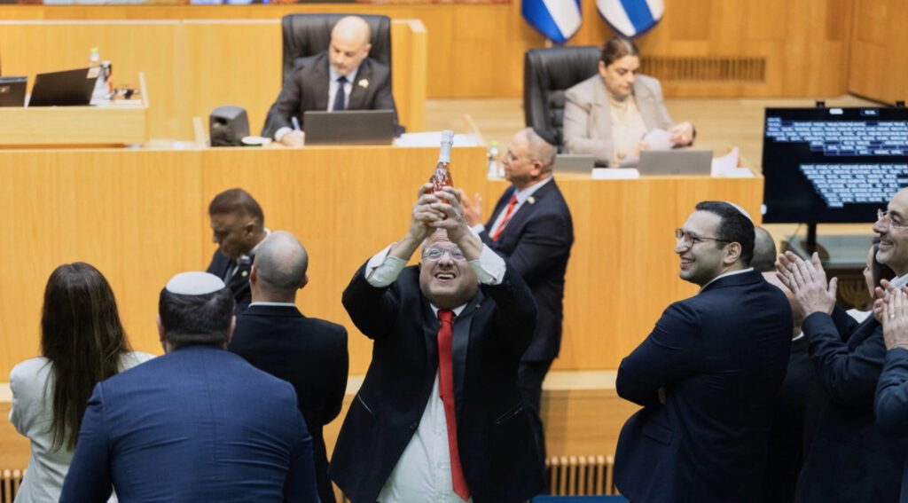 El ministro Itamar Ben-Gvir en el centro del parlamento israelí, levantando una botella con ambas manos en un gesto de festejo, rodeado de legisladores que aplauden y sonríen.