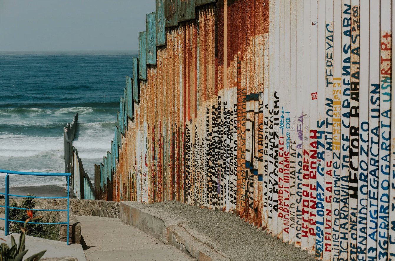Vista del muro fronterizo metálico que se extiende sobre la arena hasta el Océano Pacífico en Tijuana, cubierto de grafitis y nombres, bajo un cielo despejado.