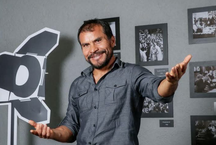 El actor Noé Hernández sonriendo con los brazos abiertos en una camisa gris, posando junto a una figura recortada de un reflector de cine y una pared gris decorada con fotografías cinematográficas en blanco y negro.