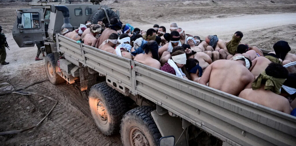 Decenas de hombres palestinos detenidos, con los ojos vendados y con el torso desnudo, son transportados hacinados en la parte trasera de un camión militar en un terreno de tierra.