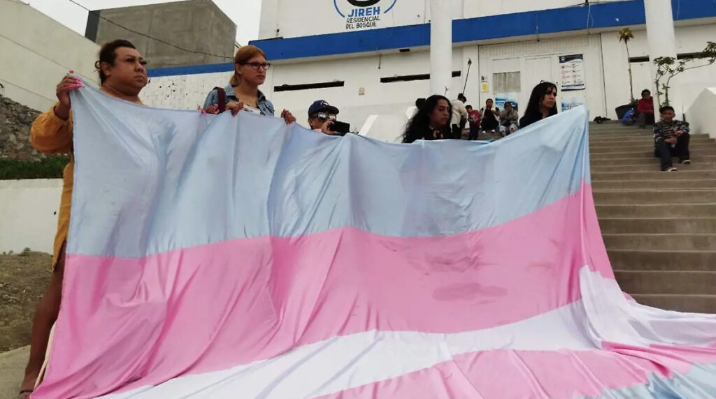 Activistas y mujeres trans sostienen una bandera gigante trans durante una protesta frente al centro Jireh Residencial del Bosque en Tijuana, denunciando violencia de la Patrulla Espiritual.