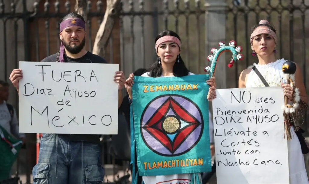 Manifestantes indígenas sostienen pancartas que dicen "Fuera Diaz Ayuso de México" y "No eres bienvenida Diaz Ayuso", junto a estandartes culturales durante la gira de la mandataria española.