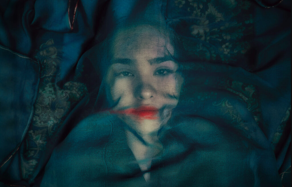 Primer plano artístico de un rostro femenino envuelto en una tela traslúcida de color azul profundo, con labios acentuados en rojo, fotograma del documental 'Un zorro bajo una luna rosa'.