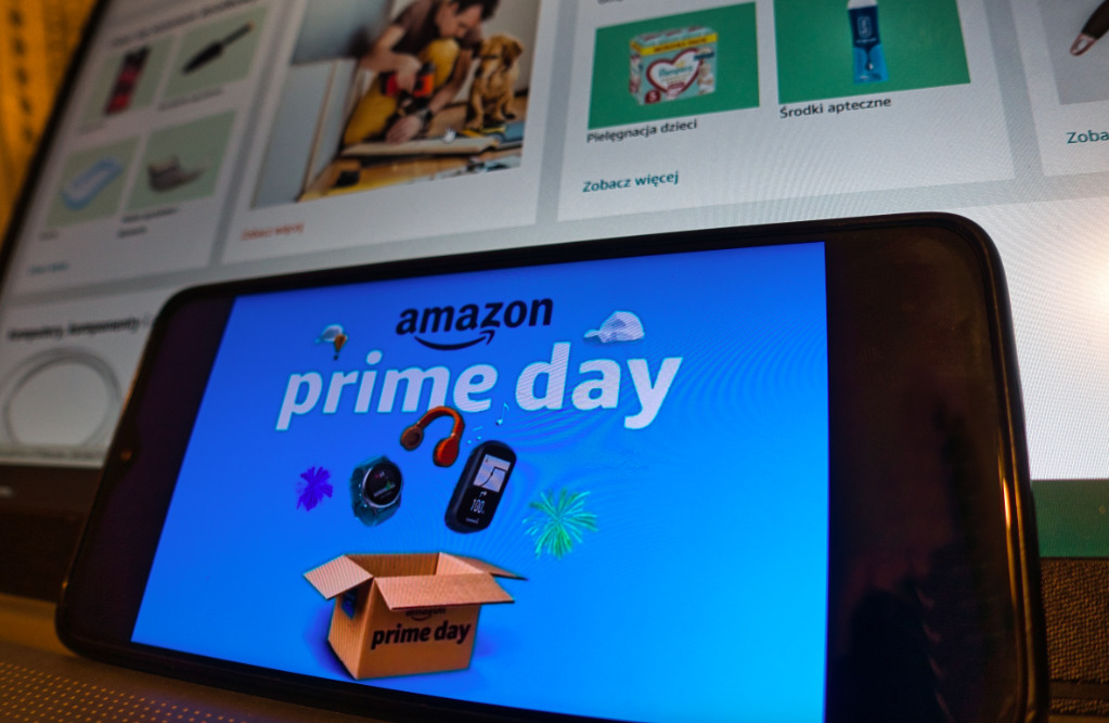 Primer plano de la pantalla de un teléfono celular que muestra el logotipo oficial de Amazon Prime Day sobre un fondo azul con gráficos de una caja de cartón abierta, audífonos y un reloj inteligente. Al fondo, de forma desenfocada, se observa la pantalla de una computadora portátil con un catálogo de productos de comercio electrónico.