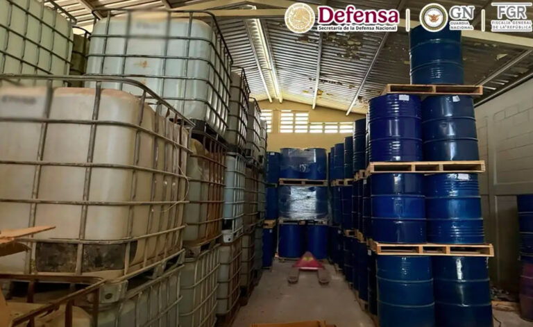 Interior de un almacén o bodega con techo de lámina lleno de contenedores industriales. A la izquierda se aprecian grandes contenedores de plástico blanco con rejillas metálicas (IBC) apilados, y a la derecha, múltiples tambos de metal azul marino colocados sobre tarimas de madera. En la parte superior derecha se observan los logotipos oficiales de Defensa, Guardia Nacional y la FGR.