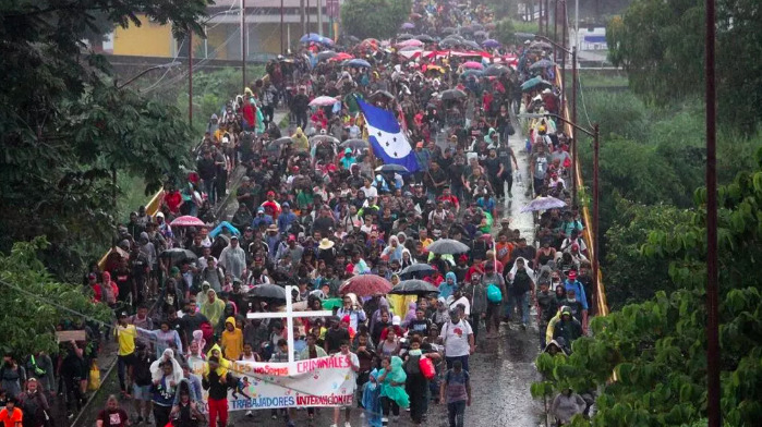 Vista elevada de una multitud masiva de migrantes caminando por una carretera bajo la lluvia en Tapachula. Llevan paraguas, impermeables, una gran bandera de Honduras azul y blanca, y una manta al frente con una cruz blanca que dice No somos criminales.
