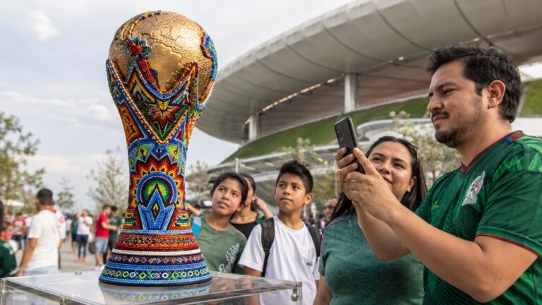 Una réplica dorada de la Copa del Mundo de la FIFA con la base y el cuerpo detalladamente decorados con chaquiras de colores en patrones tradicionales de arte huichol (wixárika). En primer plano, un hombre con playera de la selección mexicana toma una foto con su celular, mientras una mujer y dos niños observan la pieza afuera de un moderno estadio de fútbol.