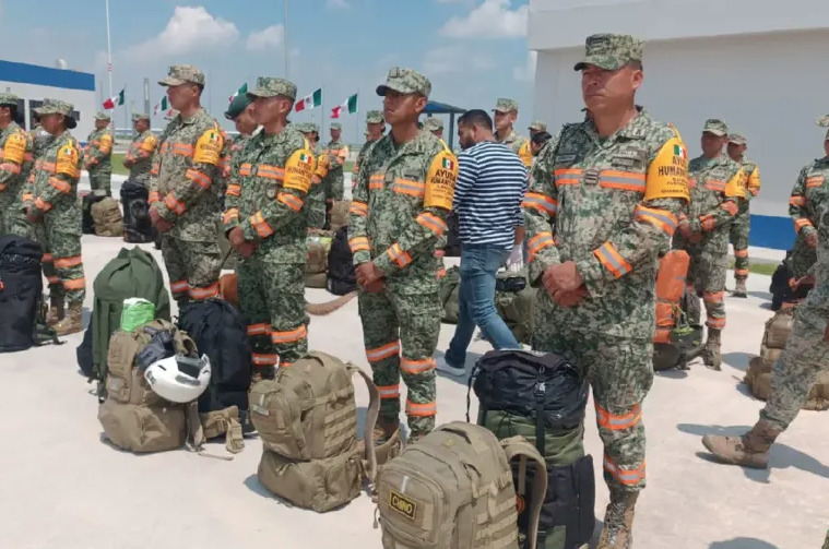 Fotografía de alta definición y gran angular de una gran formación de militares mexicanos con uniformes de camuflaje completos, franjas reflectantes naranjas y distintivos específicos de la misión, formados en filas disciplinadas sobre una extensa pista de concreto. En primer plano, numerosas mochilas militares color arena del desierto están ordenadas cuidadosamente, incluyendo una claramente marcada con la palabra "CHINO". La perspectiva más amplia revela filas de banderas mexicanas en mástiles, edificios de la base con detalles azules, vehículos de apoyo terrestre del aeropuerto y la sección delantera de un gran avión de transporte FAM C-130, todo bajo un vasto cielo azul con nubes bien definidas, lo que ilustra un importante despliegue de ayuda humanitaria.