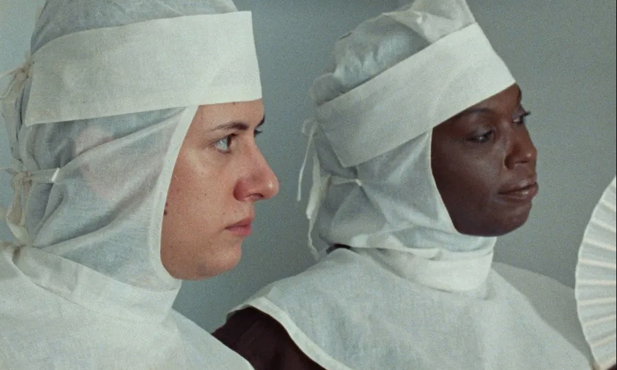 Fotograma cinematográfico de plano medio que muestra a dos mujeres sentadas de perfil hacia la derecha. Ambas visten un atuendo blanco de tela ligera que cubre su cabeza, cuello y hombros, amarrado con cintas. La mujer de la izquierda es de tez clara y mirada seria; la mujer de la derecha es de tez afrodescendiente y sostiene un abanico blanco de papel frente a su rostro en un entorno de iluminación suave.