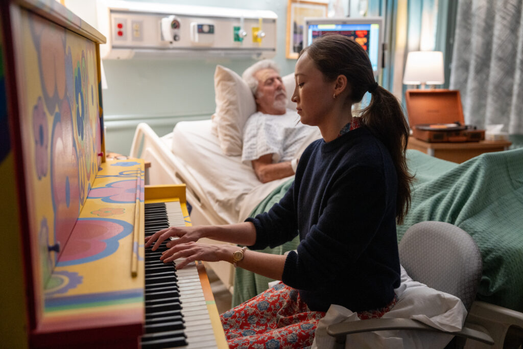 Fotograma de la película El Afinador. Una mujer con suéter oscuro toca un piano vertical pintado con flores coloridas psicodélicas, sentada de perfil en una habitación de hospital donde un hombre mayor con bata médica descansa al fondo en una cama clínica.
