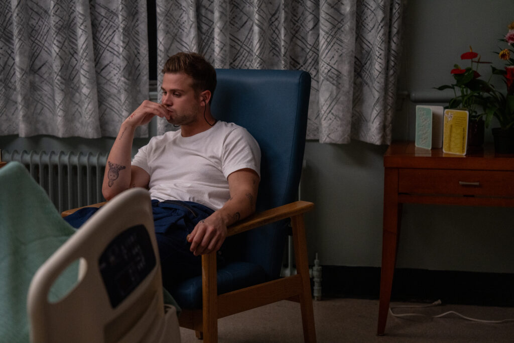 Fotograma de la película El Afinador. Un joven con camiseta blanca y tatuaje en el antebrazo derecho está sentado pensativo en un sillón azul dentro de una habitación de hospital, con una mano cerca de su rostro y una cama clínica en primer plano.