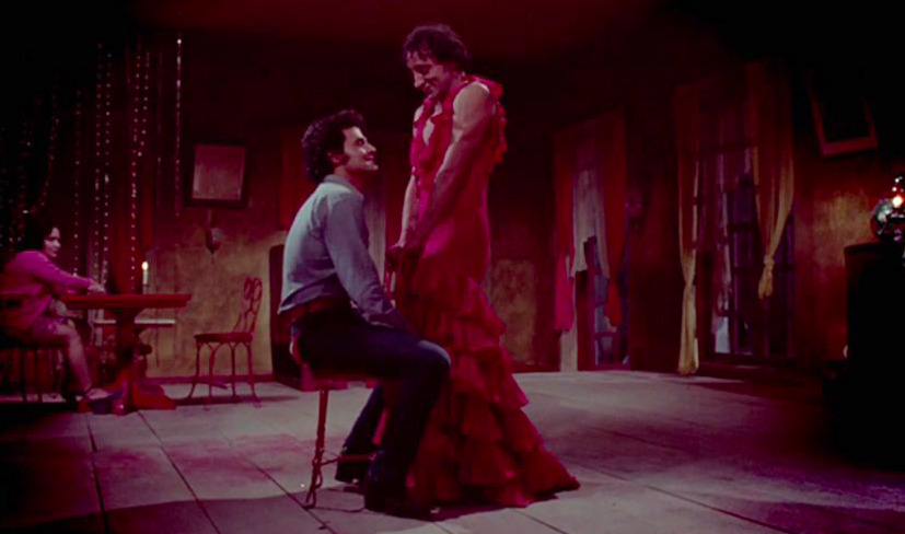 Fotograma de la película El lugar sin límites (1978). En el centro de una habitación rústica con iluminación rojiza, el actor Roberto Cobo, interpretando a La Manuela con un vestido de olanes rojo, sonríe de pie frente al actor Gonzalo Vega, en el papel de Pancho, quien está sentado de perfil mirándolo fijamente. Al fondo a la izquierda se observa a una mujer sentada junto a una mesa de madera.