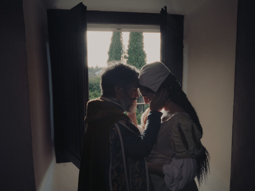 Fotograma de la película 'Magallanes' en un plano medio a contraluz frente a una ventana abierta con contraventanas de madera oscura. En la escena, un hombre con barba grisácea y vestimenta de época abraza íntimamente a una mujer que lleva un tocado blanco en la cabeza y un vestido claro con mangas abullonadas. Ambos juntan sus frentes en un momento emotivo y dramático, con una iluminación suave que entra desde el exterior mostrando árboles cipreses al fondo.