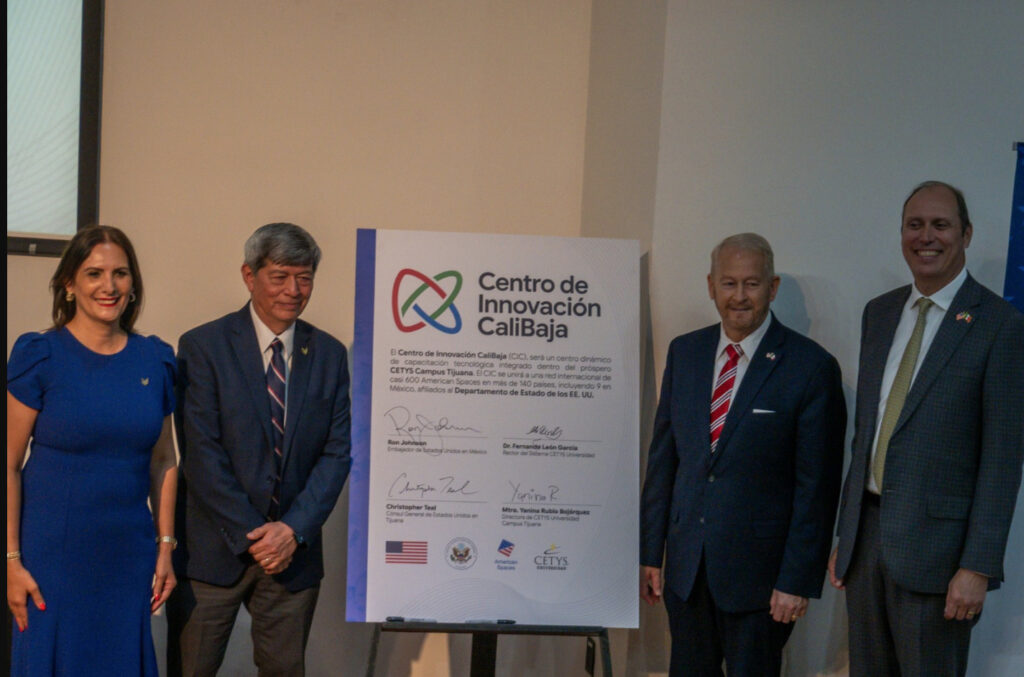 Una mujer con vestido azul rey y tres hombres en trajes formales posan de pie junto a un gran panel blanco sobre un caballete. El cartel muestra el logotipo del Centro de Innovación CaliBaja y las firmas manuscritas de autoridades como Ken Salazar, Embajador de EE. UU. en México, y el Dr. Fernando León García, Rector del Sistema CETYS Universidad, junto a logotipos oficiales de American Spaces y CETYS.
