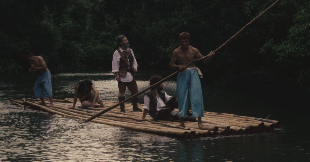 Fotograma de la película 'Magallanes' que muestra a Gael García Bernal caracterizado como Fernando de Magallanes de pie en el centro de una balsa de troncos de bambú. Viste camisa blanca con chaleco y pantalones de cuero marrón. La balsa navega por un río de aguas tranquilas rodeado de una densa selva tropical verde. Lo acompañan otras cuatro personas: dos hombres locales sin camisa con pantalones azules que guían la balsa con largas varas de madera, un hombre sentado y una persona agachada. La escena evoca un viaje de exploración en un tono cinematográfico realista y sombrío.