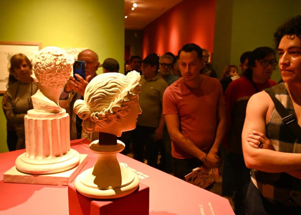 Varios visitantes en una sala de museo con iluminación cálida observando bustos de esculturas clásicas de color blanco exhibidos sobre pedestales rojos, mientras un hombre toma una fotografía con su celular al fondo.