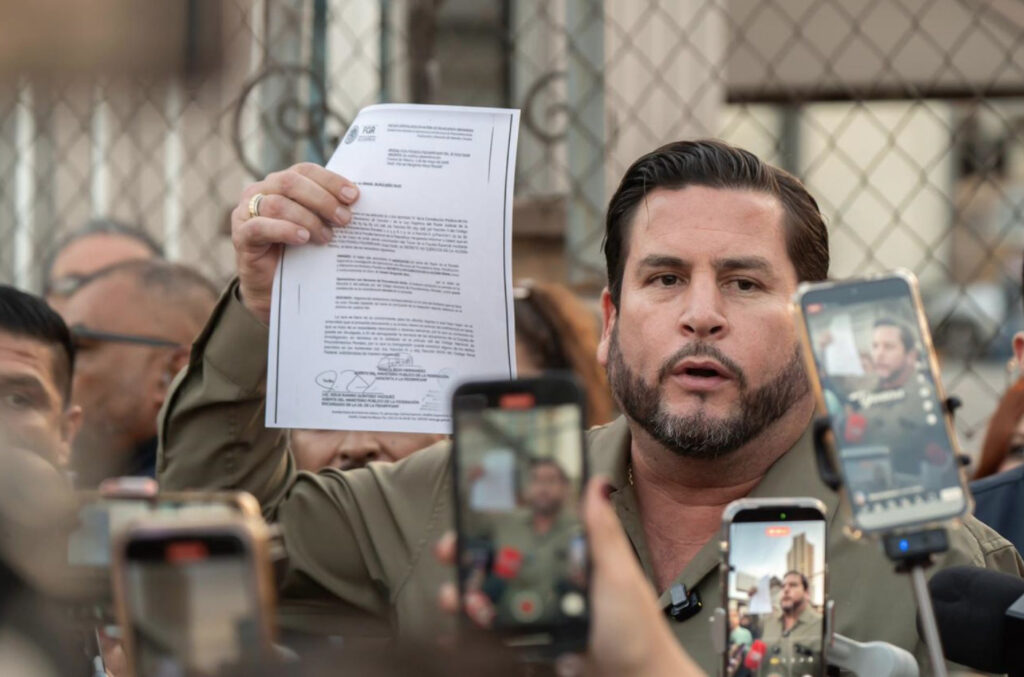 El alcalde de Tijuana, Ismael Burgueño, sostiene en alto una hoja membretada de la FGR con firmas oficiales frente a reporteros. En primer plano se aprecian varios teléfonos celulares de espaldas grabando y transmitiendo en vivo la conferencia de prensa.