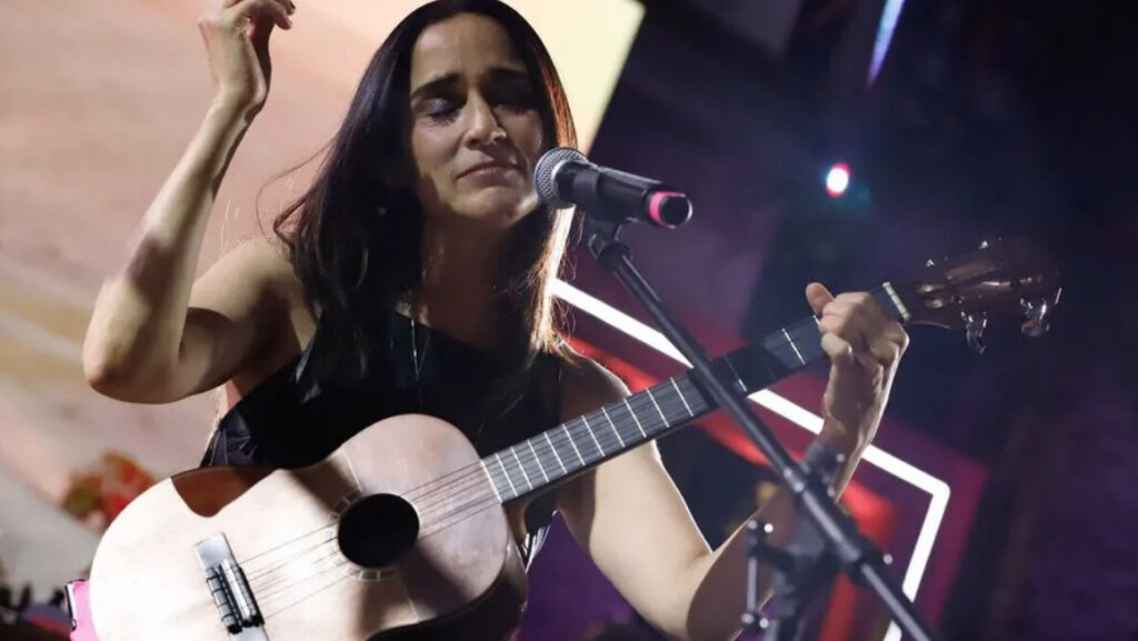 La cantante Julieta Venegas con los ojos cerrados tocando una guitarra acústica frente a un micrófono en un escenario iluminado durante la presentación de la campaña turística Baja California es para ti.