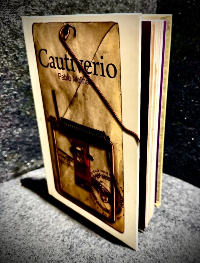 Fotografía en plano medio de la portada del libro Cautiverio de Pablo Molinet, editado por el Instituto Literario de Veracruz. El diseño de la portada, de tonos sepia y envejecidos, muestra la imagen de una trampa de alambre y madera, con un alambre real doblado que sobresale físicamente del borde del libro.