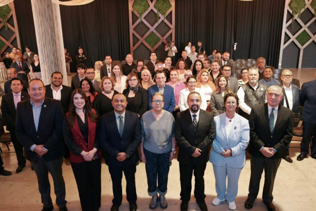 Fotografía grupal de los integrantes de la primera Mesa Directiva de la APCBC alineados al frente, vistiendo trajes formales y ropa ejecutiva, respaldados por una gran audiencia de comunicadores y periodistas de pie al fondo en un auditorio con decoraciones de madera.