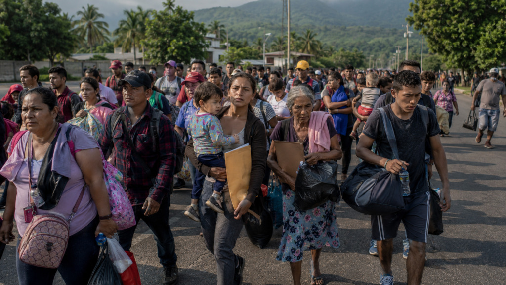 Un grupo numeroso de hombres, mujeres y niños migrantes caminan juntos por una carretera portando maletas, mochilas y carpetas con documentos, en una toma exterior de día con vegetación y cerros al fondo.