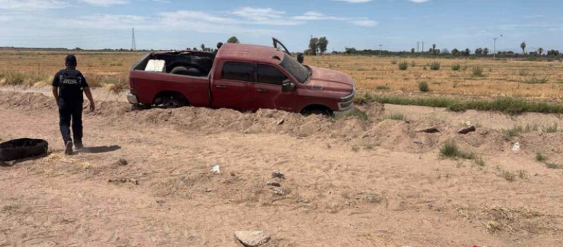 Una camioneta pick-up de cuatro puertas color rojo ocre se encuentra varada y semi-atascada en una zanja de tierra en medio de una zona rural o campo de cultivo seco. A la izquierda, un elemento policiaco uniformado de espaldas con la leyenda "Fuerza Estatal" camina hacia el vehículo bajo un cielo parcialmente despejado.