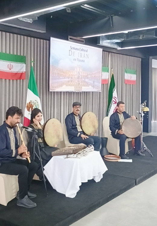 Un ensamble de músicos tradicionales iraníes sentados en un escenario interpretando instrumentos de viento y percusión como el daf. El fondo de la tarima está decorado con banderas de México e Irán, y un cartel superior que dice Semana Cultural DE IRÁN en Tijuana.