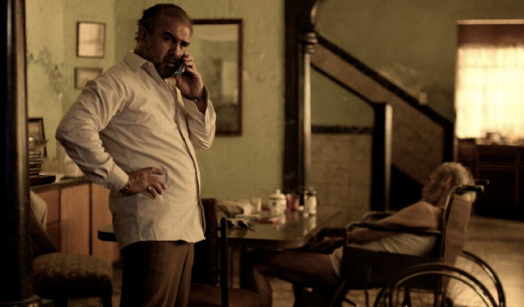 Fotograma en tonos sepia y verdosos de la película 'Tus dos muertos'. Un hombre con camisa blanca habla por teléfono celular de pie con una mano en la cintura, mientras al fondo un anciano descansa en una silla de ruedas junto a una mesa de comedor.