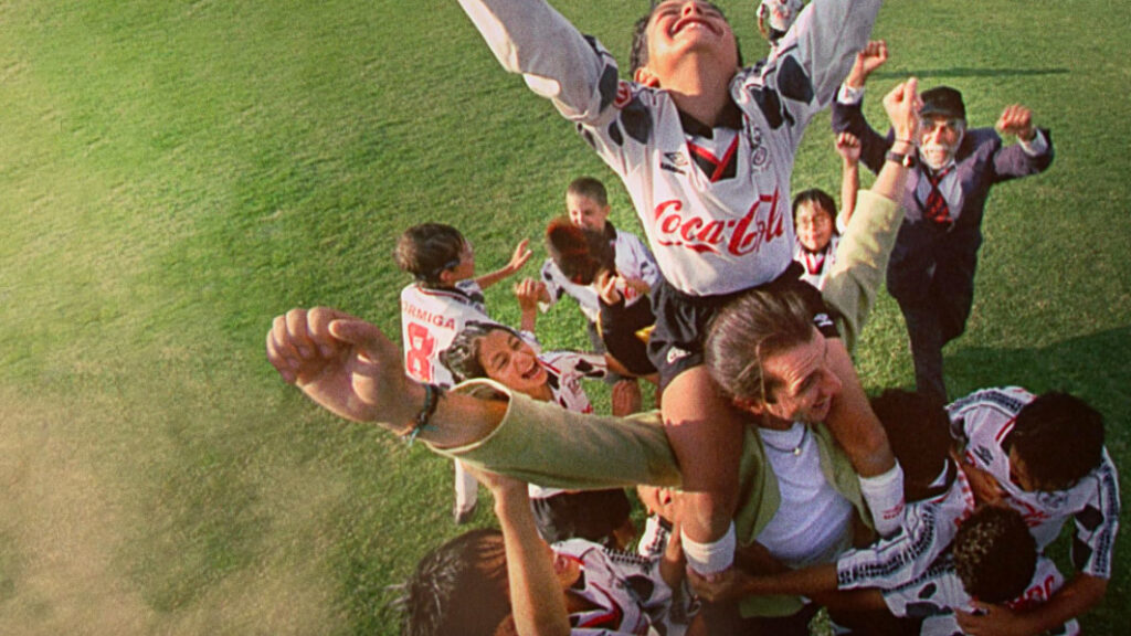 Fotograma cinematográfico de la película mexicana 'Atlético San Pancho' (2001) tomado desde un ángulo cenital elevado. Muestra una celebración de victoria en una cancha de fútbol de pasto verde. En el centro, un niño futbolista sonríe con los brazos levantados al cielo mientras festeja montado sobre los hombros de su entrenador, quien viste una playera blanca y saco verde claro. A su alrededor, otros niños del equipo con uniformes blancos y negros de la marca Umbro con el patrocinio de Coca-Cola corren y gritan de alegría. Al fondo a la derecha, un hombre mayor de barba blanca con traje oscuro y corbata corre con los puños en alto festejando.