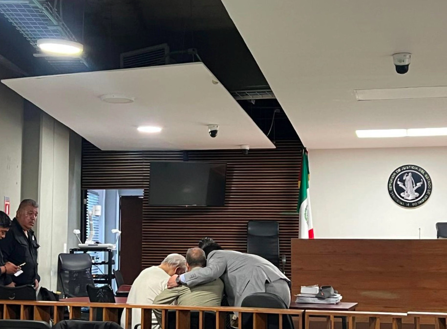 Fotografía del interior de una sala de audiencias judiciales en el estado de Morelos. En primer plano, sentados detrás de un barandal de madera, se observa de espaldas al exdirector de Pemex, Víctor Rodríguez Padilla, vistiendo ropa clara y con la cabeza inclinada hacia abajo, junto a sus abogados defensores; uno de ellos, con saco gris, le coloca un brazo sobre la espalda en señal de apoyo. Al fondo a la derecha destaca el estrado de madera del tribunal con el logotipo oficial del Tribunal Superior de Justicia del Estado de Morelos y la bandera de México. En el extremo izquierdo, un elemento de seguridad con uniforme oscuro permanece de pie vigilando la sala.