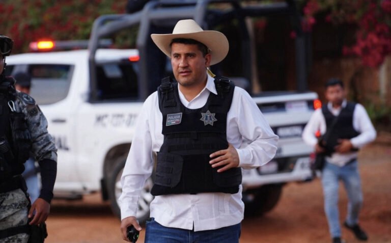 Fotografía en plano medio del exalcalde de Uruapan, Carlos Manzo, caminando en un operativo de campo. Viste una camisa blanca de manga larga, sombrero claro de ala ancha y un chaleco táctico negro con la insignia de la Policía Municipal. En su mano derecha sostiene un equipo de radiocomunicación, mientras de fondo se aprecia una camioneta patrulla blanca de la Policía Municipal con torretas encendidas y elementos de seguridad custodiando el área.