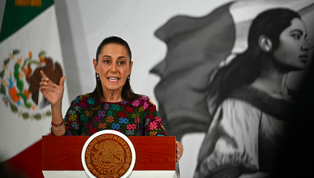 Fotografía de la presidenta de México, Claudia Sheinbaum, de pie detrás de un podio de madera con el Escudo Nacional que dice 'Estados Unidos Mexicanos'. Viste una blusa negra bordada con flores de vivos colores (rosa, azul, verde y amarillo). Se encuentra gesticulando con la mano derecha levantada mientras habla con expresión firme durante una conferencia de prensa. De fondo a la izquierda se aprecia la bandera de México y a la derecha una ilustración en blanco y negro de la herencia mexicana con una mujer patria.