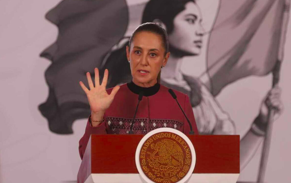 Fotografía en plano medio de la presidenta Claudia Sheinbaum Pardo vistiendo una prenda color guinda con bordados tradicionales en el pecho y cuello alto negro. Aparece de pie detrás del pódium presidencial con el Escudo Nacional, levantando la mano derecha con los dedos extendidos mientras habla frente a dos micrófonos. De fondo se aprecia la ilustración institucional en tonos grises.