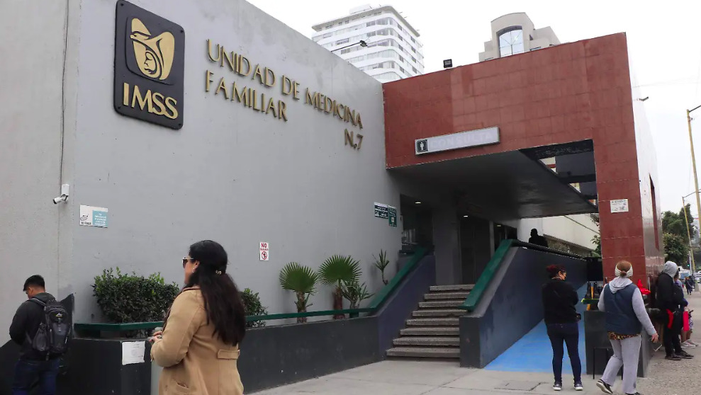 Fotografía en plano general que muestra la entrada exterior de la Unidad de Medicina Familiar Número 7 del IMSS. El edificio presenta una pared principal gris con letras metálicas doradas que dicen 'UNIDAD DE MEDICINA FAMILIAR N. 7' junto al logotipo oficial negro y dorado del IMSS. A la derecha, una estructura de fachada marrón enmarca unas escaleras de concreto y una rampa azul con barandales verdes que conducen al área de consulta. Varias personas caminan y esperan afuera de las instalaciones bajo una luz de día difusa, con edificios urbanos de fondo.