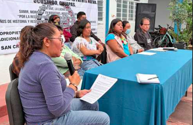 Fotografía en plano medio que muestra una conferencia de prensa al aire libre en Chiapas. En primer plano a la izquierda, una mujer de perfil con lentes y cabello recogido en una coleta viste una playera gris de manga larga y sostiene un micrófono en su mano derecha mientras lee un documento impreso. Detrás de una larga mesa cubierta con un mantel azul, se encuentran sentadas otras cinco personas escuchando con atención, entre ellas mujeres indígenas con blusas tradicionales de bordados coloridos. Al fondo se observa parte de una lona blanca con consignas de protesta impresas en letras negras y una bicicleta estacionada.