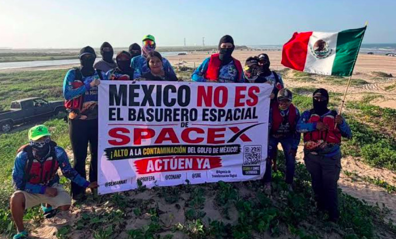 Fotografía en plano general de integrantes de la organización Conibio Global posando en la vegetación costera de Playa Bagdad en Matamoros, Tamaulipas. El grupo sostiene una pancarta blanca con letras grandes que dice 'MÉXICO NO ES EL BASURERO ESPACIAL DE SPACEX ¡ALTO A LA CONTAMINACIÓN DEL GOLFO DE MÉXICO! ACTÚEN YA'. Un participante a la derecha sostiene la bandera nacional de México, mientras otros llevan cubrebocas y ropa de protección en un entorno de dunas y mar al fondo.