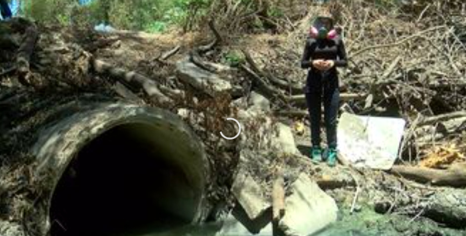 Fotografía en plano general de una zona de drenaje pluvial rodeada de ramas secas, escombros de concreto y desechos. A la izquierda destaca la gran embocadura circular de una tubería de concreto por la que fluyen aguas grises. A la derecha, una persona viste ropa oscura, calzado de seguridad azul y una máscara respiratoria de protección con filtros rosados mientras observa el área contaminada.