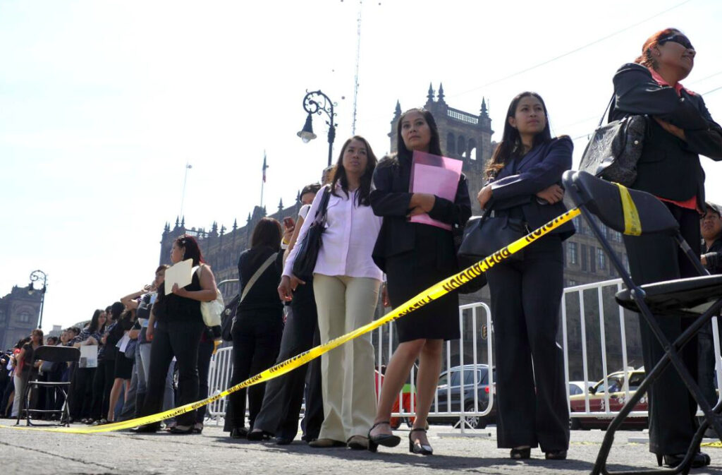 Fotografía en plano general desde un ángulo contrapicado que muestra a una larga fila de personas, principalmente mujeres vestidas con ropa formal de oficina y portando carpetas con currículums, esperando en la vía pública delimitada por vallas metálicas y una cinta amarilla de precaución. Al fondo se aprecian edificios históricos bajo un cielo despejado.