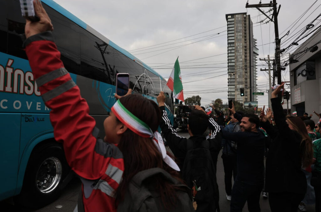 Fotografía a nivel de calle que muestra a un grupo de aficionados en Tijuana despidiendo con entusiasmo a la selección de fútbol de Irán tras el Mundial 2026. En primer plano, una joven de espaldas con una cinta tricolor en la cabeza levanta su teléfono celular hacia un gran autobús de pasajeros azul y gris en movimiento. Al fondo, la multitud levanta los brazos, ondea una bandera de Irán y sonríe en una calle urbana con postes de luz, cables y un rascacielos moderno bajo un cielo nublado.