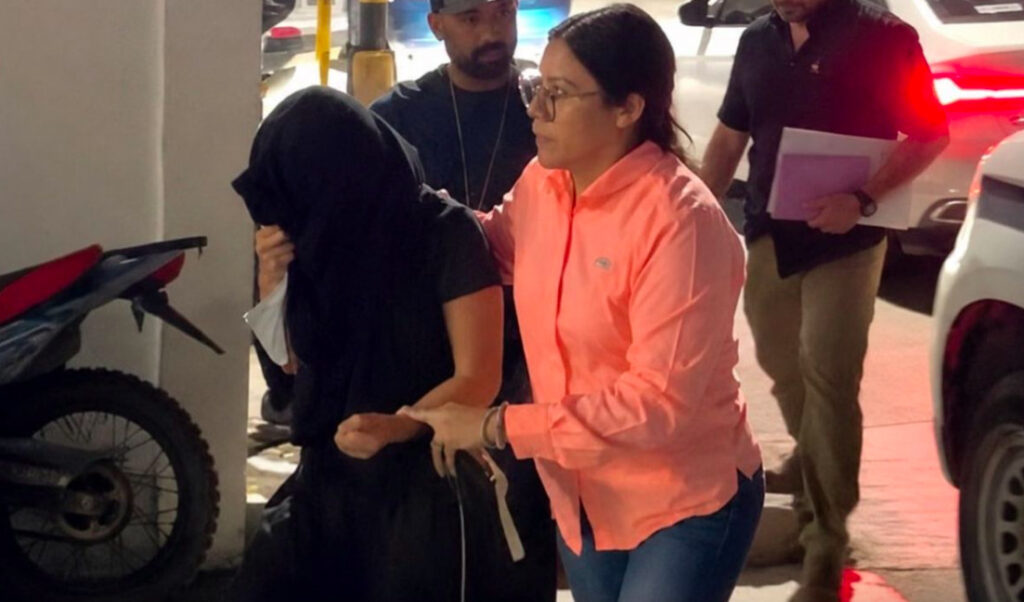Fotografía en plano general medio que muestra la custodia y traslado de una detenida durante la noche. A la izquierda, una mujer viste ropa oscura y lleva la cabeza completamente cubierta con una tela negra para ocultar su rostro mientras es escoltada. A su lado, una agente o investigadora con lentes y camisa de manga larga color salmón o naranja claro la sujeta del brazo. Al fondo, un hombre con gorra negra y otro sujeto con camisa oscura y un folder morado en la mano caminan cerca de vehículos estacionados.