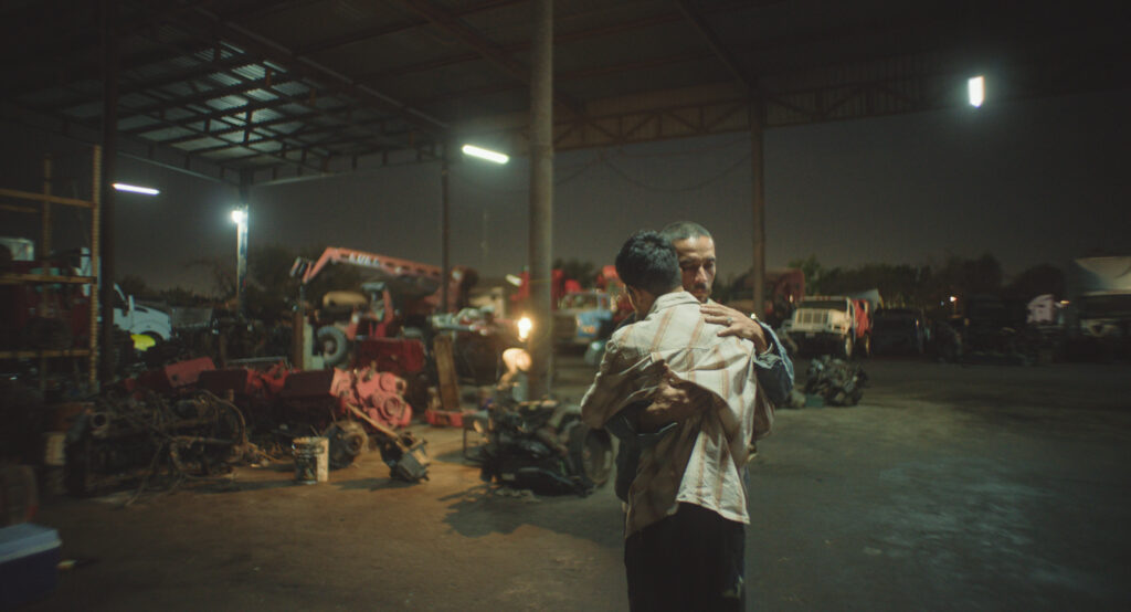 Fotograma cinematográfico de la película 'En el camino' que muestra a dos hombres abrazándose emotivamente en el centro de un gran taller mecánico o deshuesadero de vehículos techado durante la noche. El hombre en primer plano está de espaldas vistiendo una camisa a cuadros clara, mientras que el otro hombre lo sujeta firmemente con una expresión de dolor o consuelo en su rostro. El entorno industrial está iluminado por lámparas fluorescentes colgadas del techo y luces distantes, dejando ver de fondo herramientas pesadas, motores desarmados, camiones y una grúa roja.