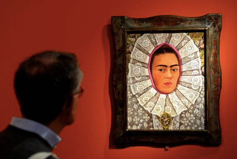 Fotografía que muestra en primer plano y desenfocado a un hombre de espaldas observando la pintura 'Autorretrato con medallón' (1948) de Frida Kahlo. La obra, colgada en una pared de color rojo terracota, está enmarcada en un marco de madera oscura labrada. El óleo muestra el rostro de Frida Kahlo con su característica uniceja y expresión seria, envuelto en un resplandor rosa y un gran tocado o cuello de encaje blanco con patrones florales detallados, portando un medallón dorado en la parte inferior.