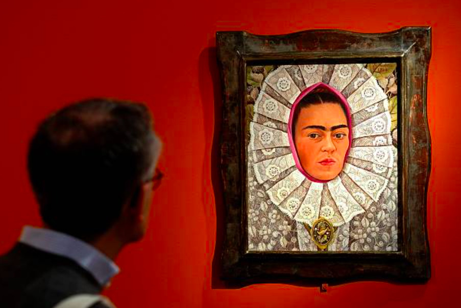 Fotografía que muestra en primer plano y desenfocado a un hombre de espaldas observando la pintura 'Autorretrato con medallón' (1948) de Frida Kahlo. La obra, colgada en una pared de color rojo terracota, está enmarcada en un marco de madera oscura labrada. El óleo muestra el rostro de Frida Kahlo con su característica uniceja y expresión seria, envuelto en un resplandor rosa y un gran tocado o cuello de encaje blanco con patrones florales detallados, portando un medallón dorado en la parte inferior.