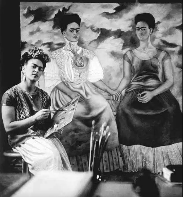 Fotografía histórica en blanco y negro de la artista mexicana Frida Kahlo sentada en su estudio. Aparece del lado izquierdo, vistiendo un huipil bordado tradicional, falda clara y flores en el cabello, mientras sostiene un pincel y una paleta de pintor con mezclas de óleo. A su derecha se encuentra su gran cuadro terminado 'Las dos Fridas' (1939), que retrata a dos versiones de sí misma sentadas tomadas de la mano, una vestida con un traje victoriano blanco con el corazón expuesto y la otra con un vestido de tehuana oscuro. En primer plano inferior se observa un frasco con múltiples pinceles.