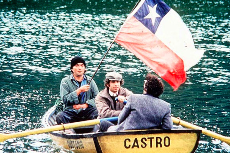 Fotograma cinematográfico de la película chilena 'Historias de fútbol' (1997) de Andrés Wood. En la escena, tres hombres navegan sobre el agua en un pequeño bote de remos de madera de color amarillo que lleva pintada la palabra 'CASTRO' en la popa. El hombre situado a la izquierda, vestido con chamarra gris y gorro negro, sostiene firmemente una gran bandera de Chile que ondea al viento. Al centro, otro hombre con boina y abrigo claro sujeta uno de los remos, mientras conversa con un tercer pasajero de espaldas que viste un saco gris texturizado.