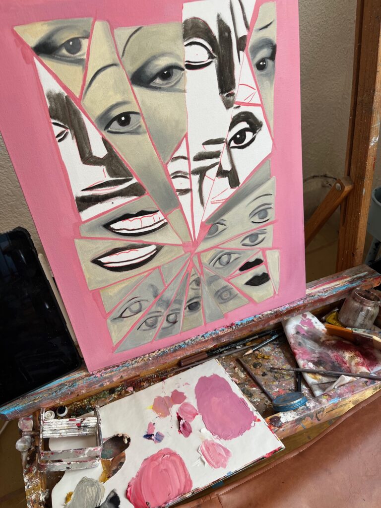 Fotografía del proceso de pintura en el estudio de la artista mexicana Mónica Loya para la identidad visual del Festival de Cine Alemán 2026. Sobre un caballete de madera manchado de pintura, se observa un lienzo con fondo rosa que muestra una ilustración al óleo de rostros, ojos y bocas fragmentados en formas geométricas inspiradas en el expresionismo alemán y la estética punk. En la parte inferior, sobre la repisa del caballete, se aprecian pinceles, una paleta con mezclas de pintura acrílica rosa y blanca, trapos de taller y un teléfono celular.