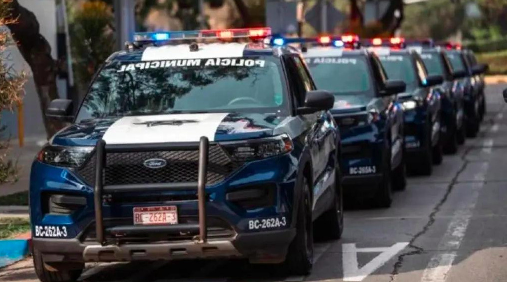Fotografía en plano general medio de una fila de patrullas SUV Ford de la Policía Municipal de Tijuana estacionadas en convoy sobre una calle urbana. En primer plano destaca la parte frontal de una camioneta azul con blanco equipada con tumba-burros, luces de emergencia rojas y azules encendidas en la torreta y número de identificación BC-262A-3, seguida por varias unidades alineadas hacia el fondo.